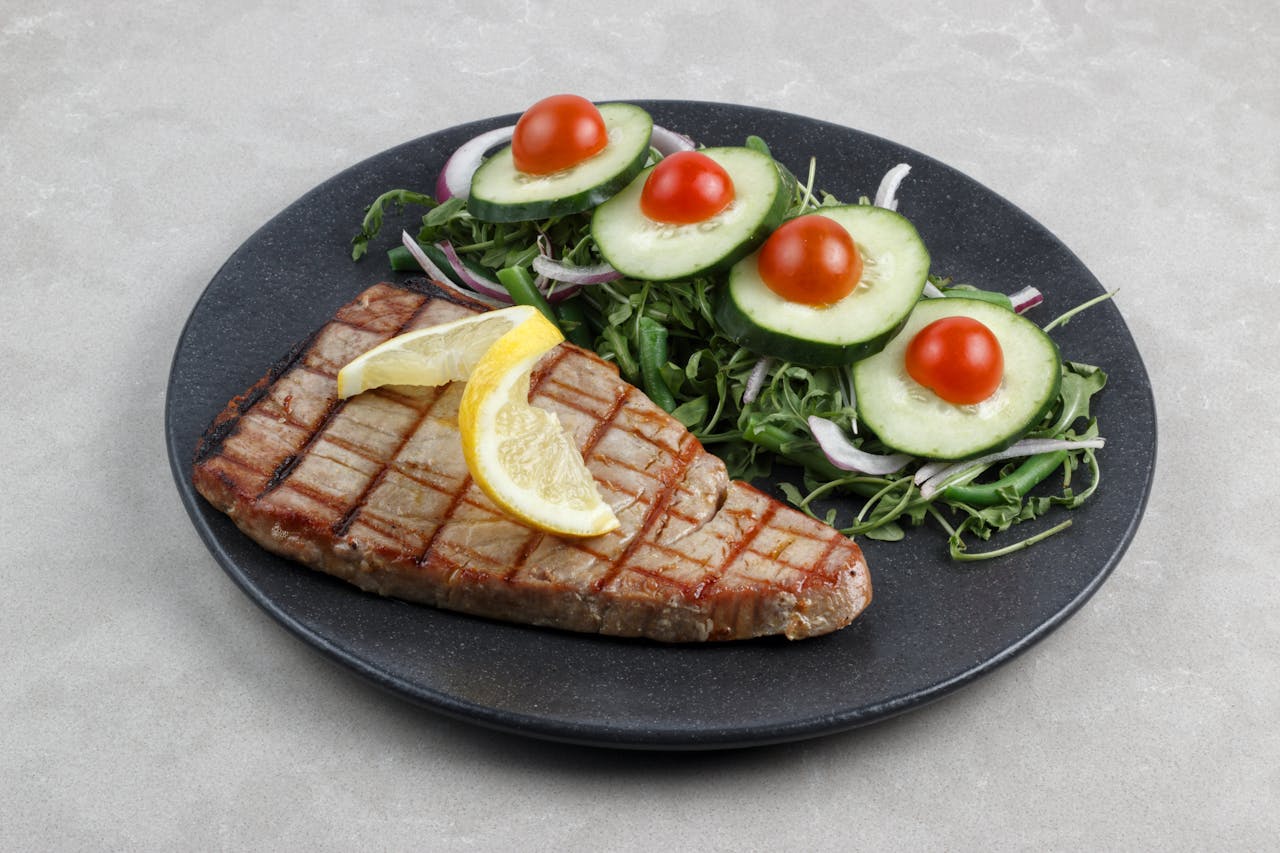 gallery-1 A grilled tuna steak topped with lemon slices served with a fresh cucumber and tomato salad on a dark plate.