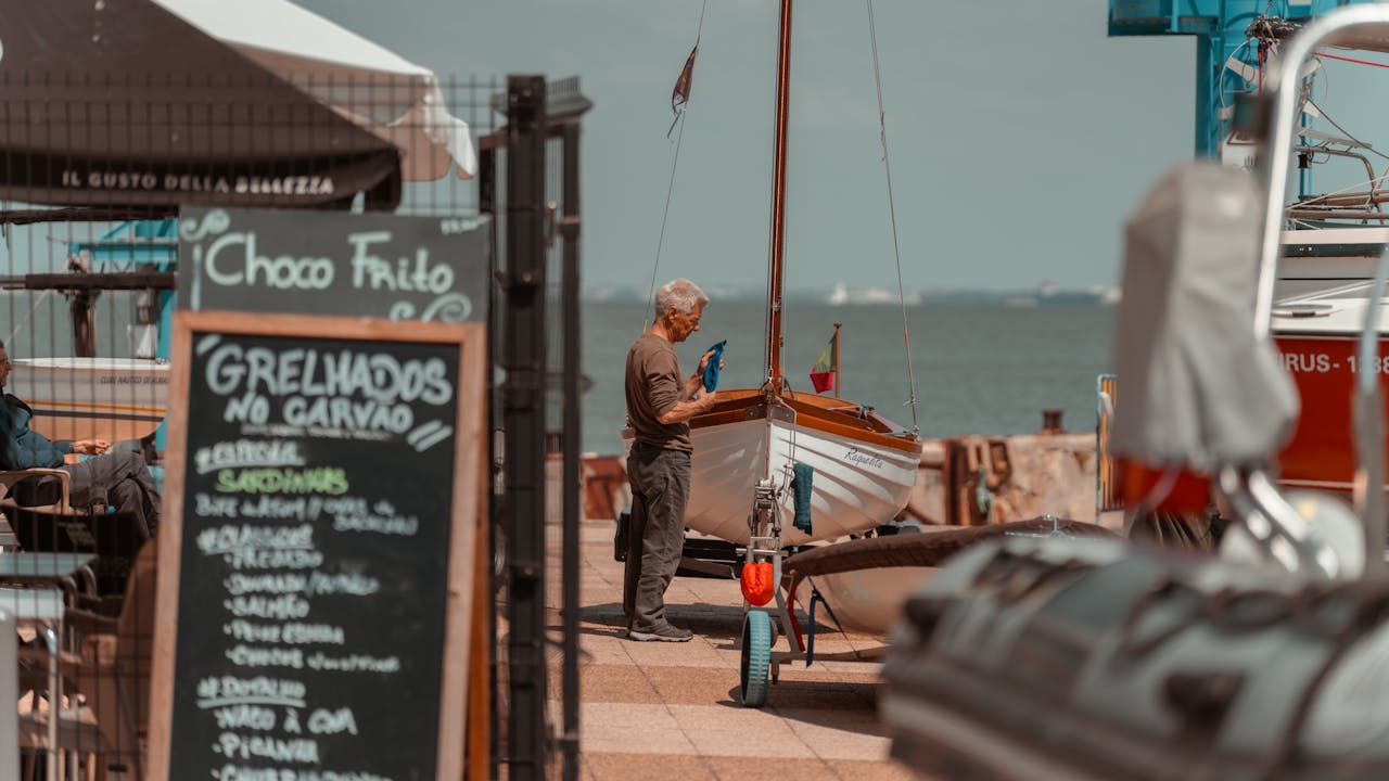 gallery-2 Elderly man inspecting a boat at a Lisbon dockside cafe with a scenic river view.