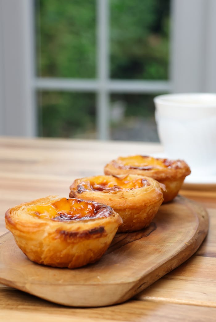 gallery-6 Close-up of three Portuguese Pastel de Nata on a wooden board indoors.