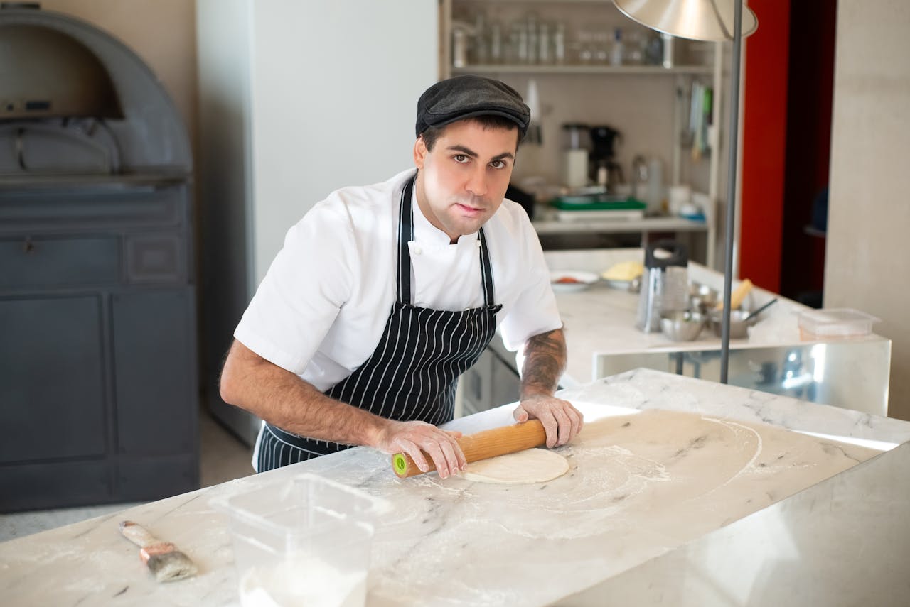 services-03 Chef in a professional kitchen rolling dough, wearing an apron and cap, perfecting his craft.