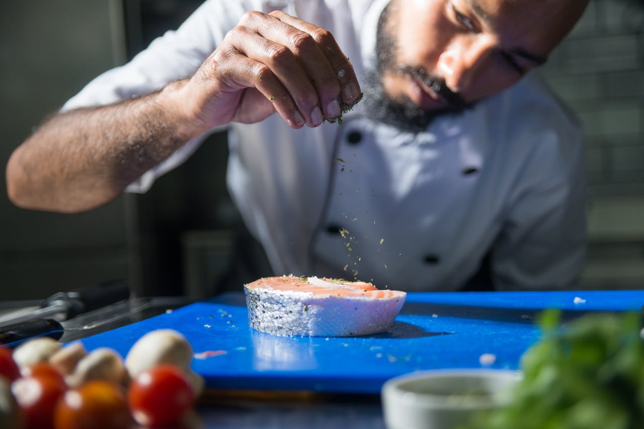 A focused chef seasoning a salmon fillet with herbs on a blue cutting board in a professional kitchen.